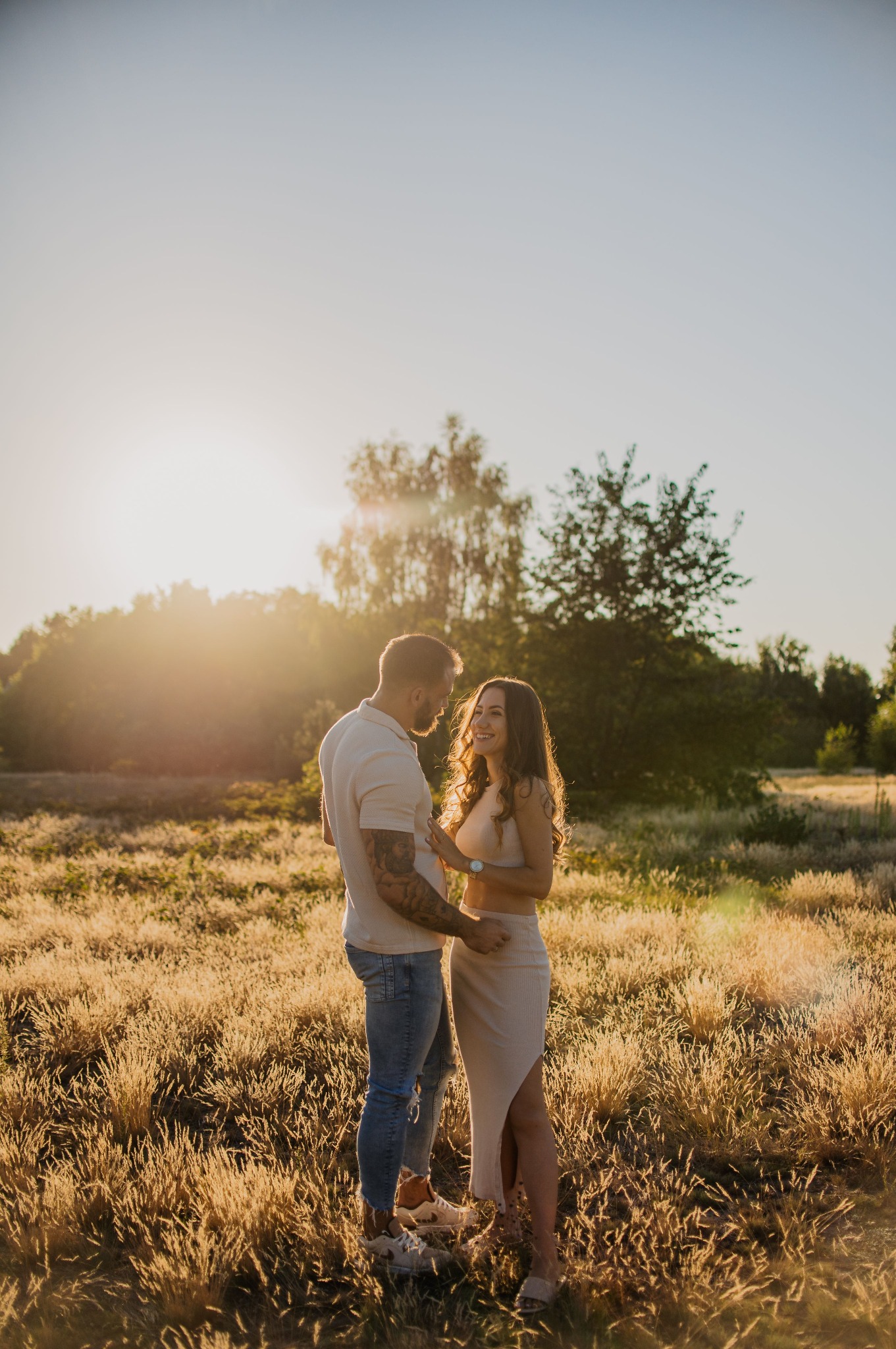Para narzeczonych w objęciach na tle łąki oświetlonej zachodzącym słońcem, mężczyzna z tatuażami na ramieniu, kobieta w beżowej sukience z rozcięciem.