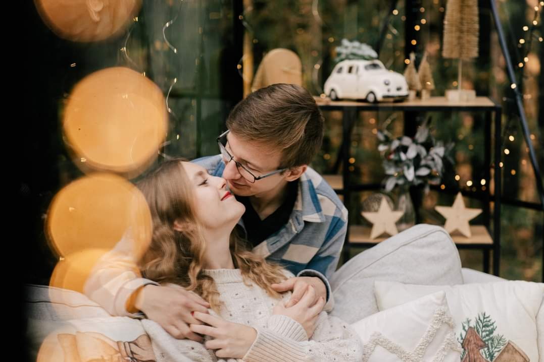 Zakochana para w objęciach na tle świątecznej dekoracji z lampkami i małym modelem samochodu z choinką na dachu.