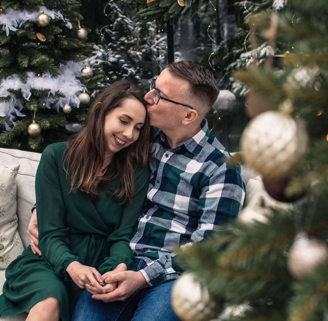 Zakochana para siedząca na kanapie, mężczyzna całuje kobietę w czoło na tle choinek z ozdobami.