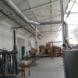 Instalacja wentylacyjna z rurami w hali produkcyjnej, widoczne maszyny i palety. Oświetlenie górne, białe ściany i sufit. Układ rur nad maszyną.