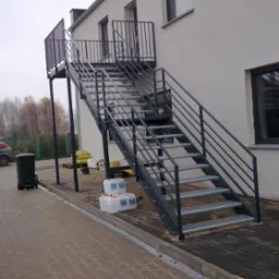 Zewnętrzne schody metalowe z balustradą, prowadzące do budynku. Widoczne elementy konstrukcyjne, siatkowe stopnie i szara elewacja budynku w tle. Ujęcie z dołu.