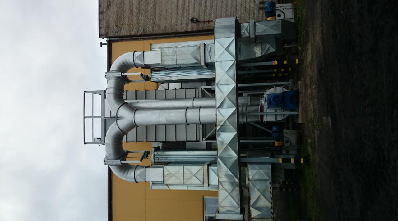 Kompleksowa instalacja wentylacyjna z rurami i wentylatorami na zewnątrz budynku, widoczna z dołu. Stalowe elementy systemu na tle żółtej ściany.