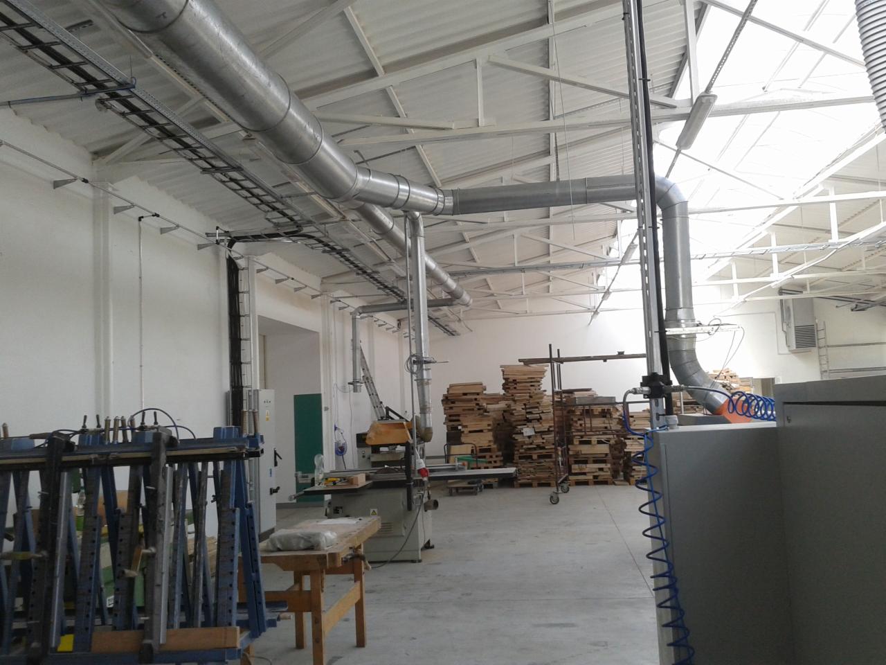 Instalacja wentylacyjna z rurami w hali produkcyjnej, widoczne maszyny i palety. Oświetlenie górne, białe ściany i sufit. Układ rur nad maszyną.