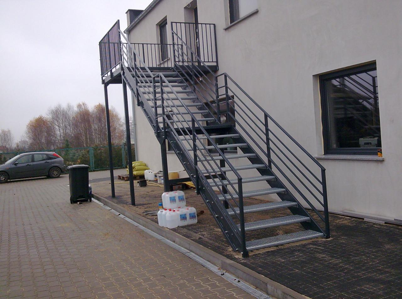 Zewnętrzne schody metalowe z balustradą, prowadzące do budynku. Widoczne elementy konstrukcyjne, siatkowe stopnie i szara elewacja budynku w tle. Ujęcie z dołu.