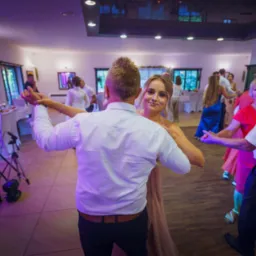 Para młoda tańczy podczas wesela, panna młoda uśmiecha się do kamery, widoczni goście i sprzęt nagłośnieniowy.