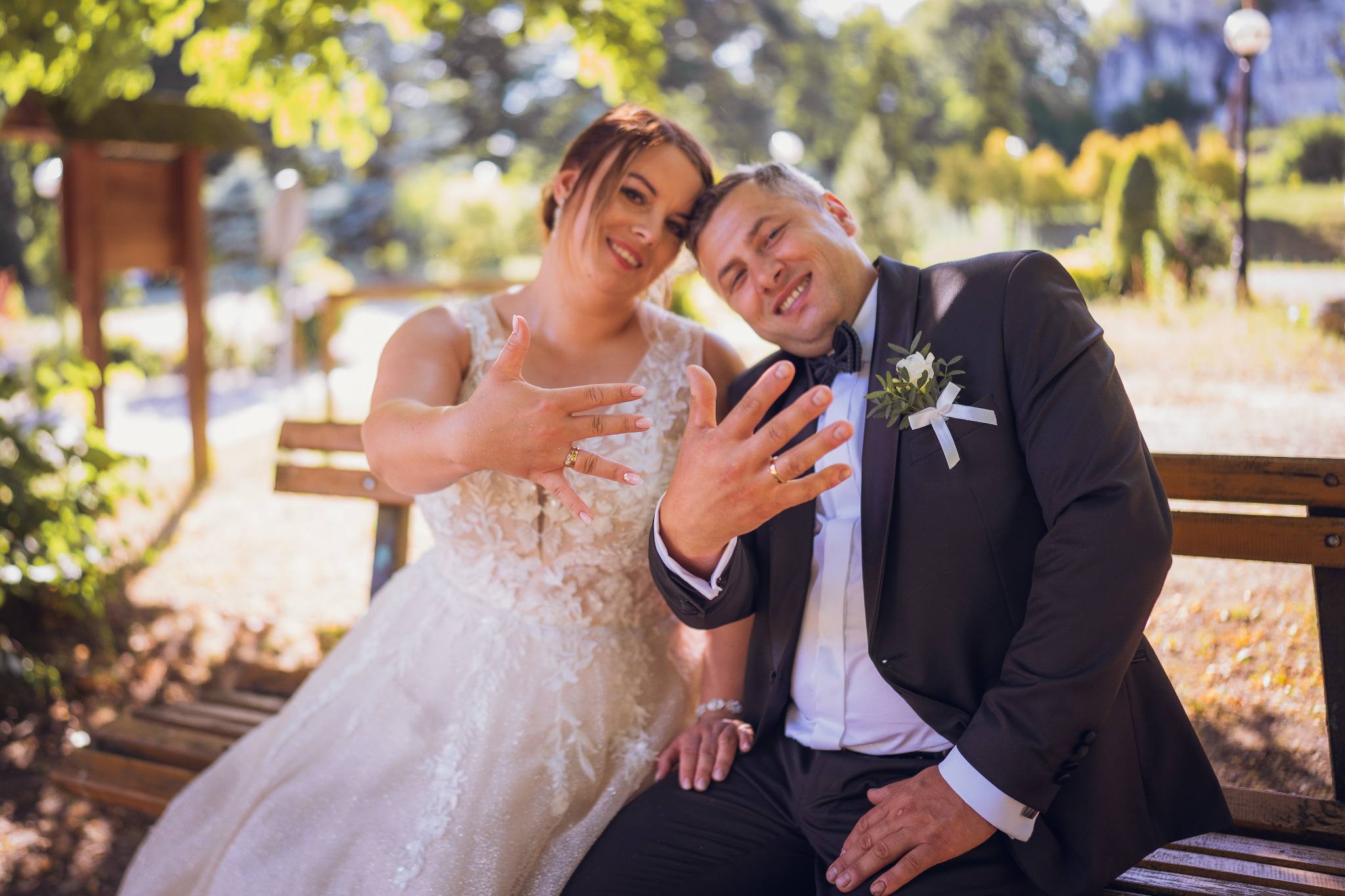 Uśmiechnięta para młoda siedzi na ławce w parku, eksponując obrączki ślubne na tle zieleni.