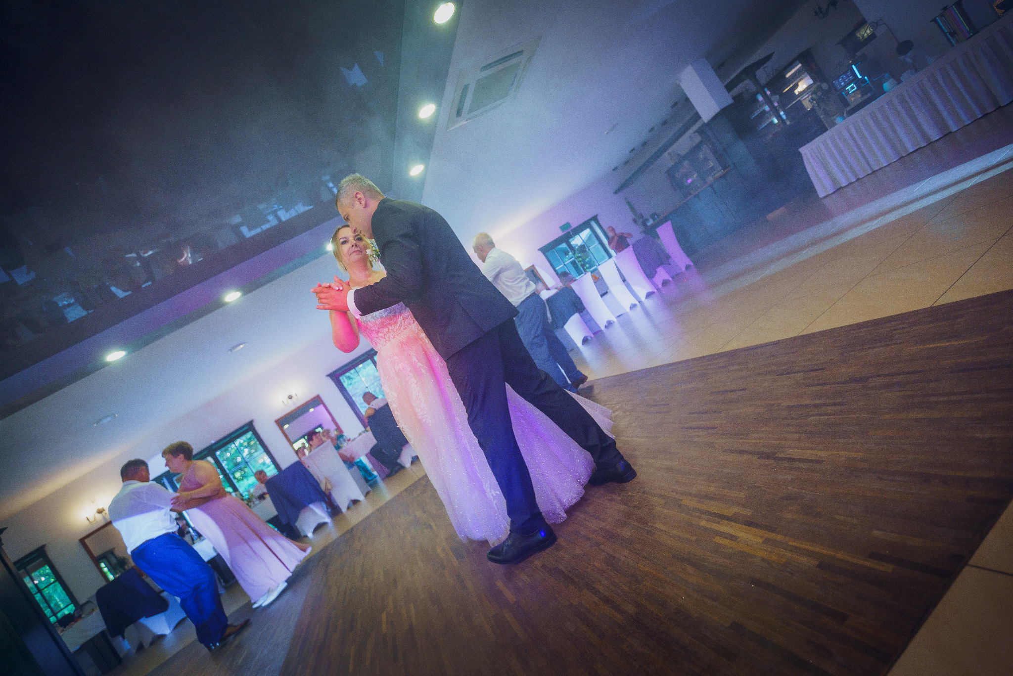 Para młoda tańczy pierwszy taniec na sali weselnej, oświetlonej fioletowymi światłami, z gośćmi siedzącymi przy stołach w tle.