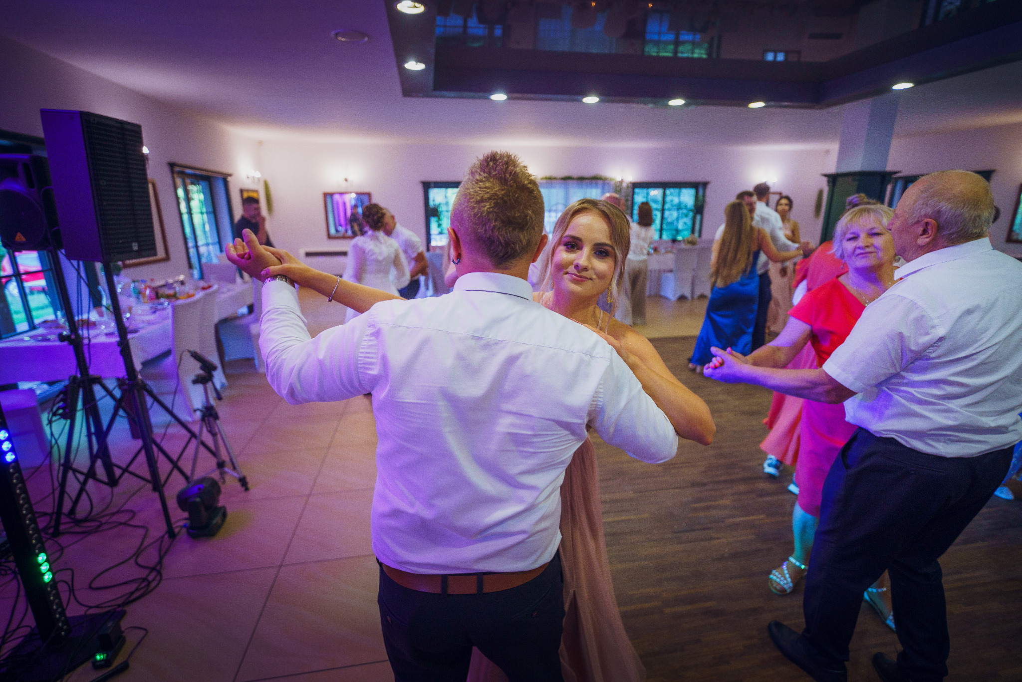 Para młoda tańczy podczas wesela, panna młoda uśmiecha się do kamery, widoczni goście i sprzęt nagłośnieniowy.