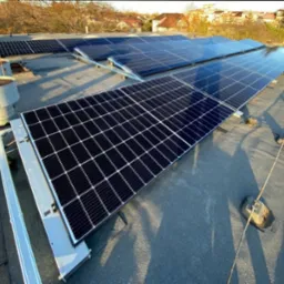 Rząd paneli słonecznych na płaskim dachu budynku mieszkalnego, widoczne kominy wentylacyjne i okoliczne domy w tle.