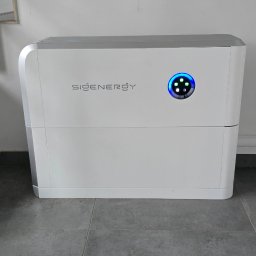 NATUR ENERGY - Biały, nowoczesny magazyn energii SiGENERGY z niebieskim wyświetlaczem, umieszczony na szarej podłodze w pobliżu schodów.