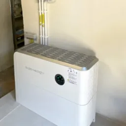 Biały magazyn energii SiCenergy w minimalistycznym wnętrzu, widoczne przewody elektryczne i rozdzielnica PV-DC na ścianie.