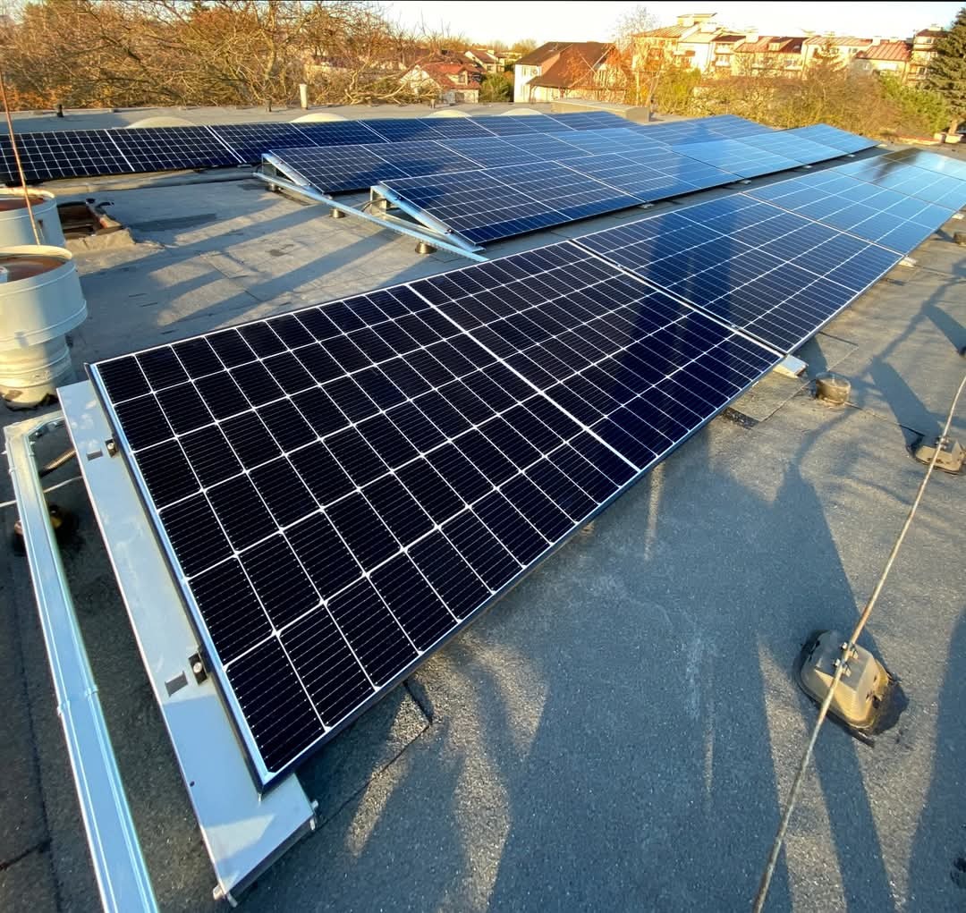 Rząd paneli słonecznych na płaskim dachu budynku mieszkalnego, widoczne kominy wentylacyjne i okoliczne domy w tle.