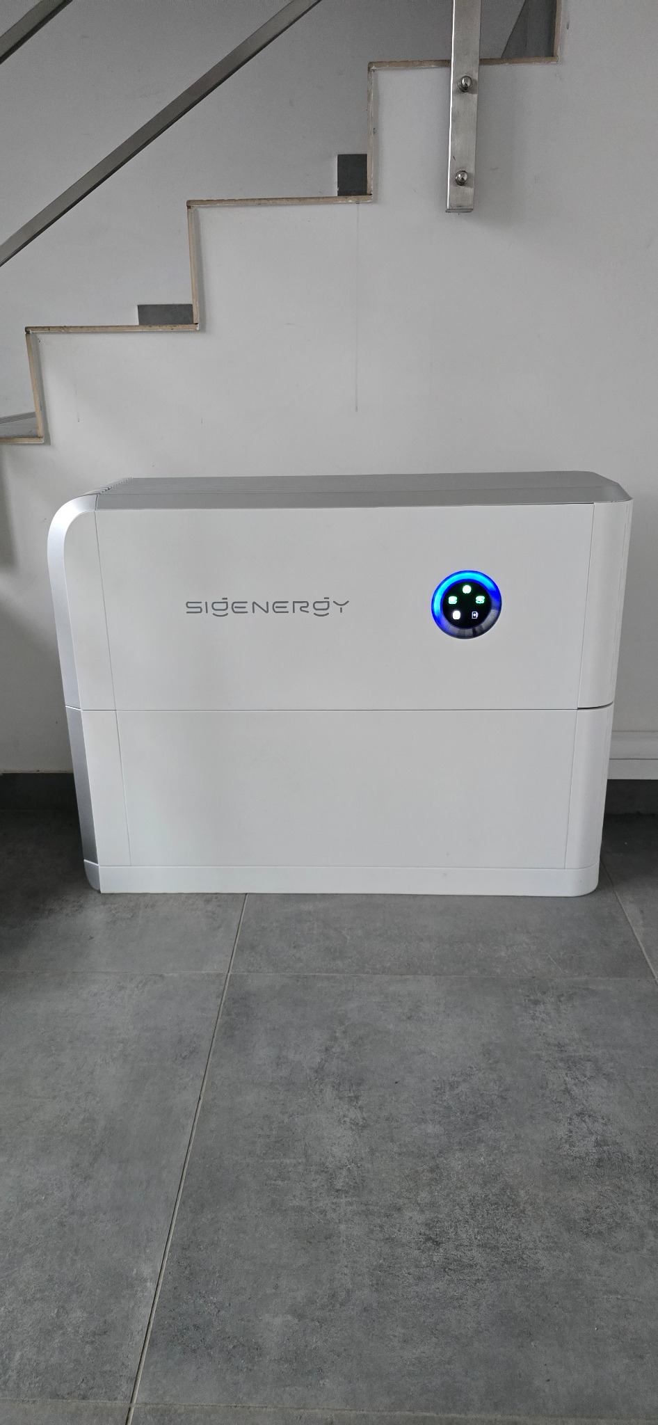 Biały, nowoczesny magazyn energii SiGENERGY z niebieskim wyświetlaczem, umieszczony na szarej podłodze w pobliżu schodów.