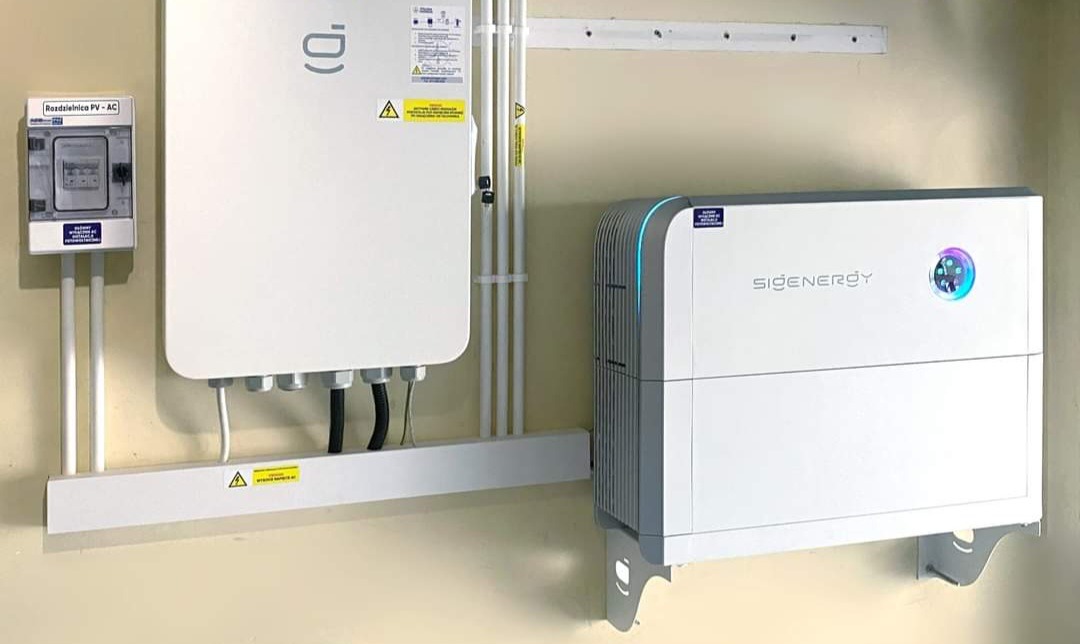 Biała instalacja fotowoltaiczna z magazynem energii SIGENERGY zamontowana na beżowej ścianie wewnątrz budynku, widoczna skrzynka rozdzielcza PV-AC i okablowanie w białych listwach.