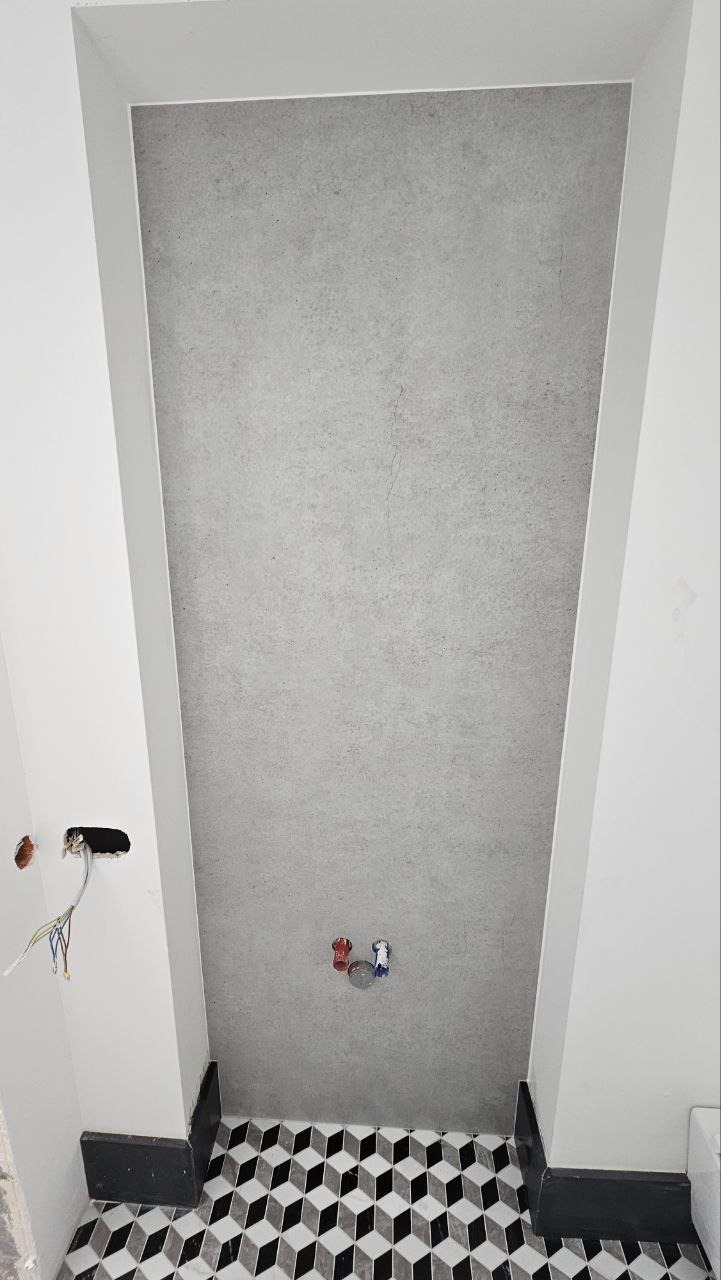 Ściana łazienki pokryta szarym tynkiem dekoracyjnym, widoczne wystające rury instalacji wodnej, fragment ściany z puszką elektryczną i wystającymi przewodami oraz podłoga wyłożona czarno-białą...