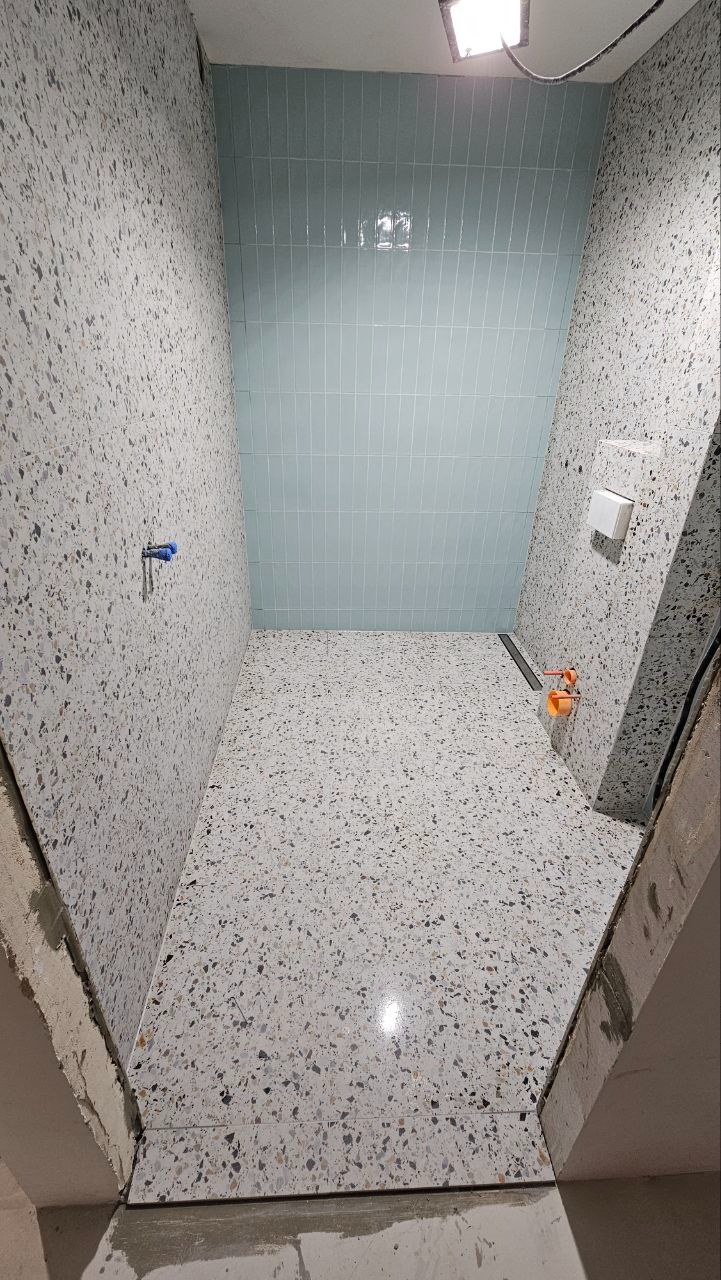 Wnętrze łazienki w trakcie prac wykończeniowych: ściany i podłoga wyłożone płytkami imitującymi lastryko, jasnoniebieskie płytki na ścianie pod prysznicem, widoczne instalacje wodne i odpływ liniowy.