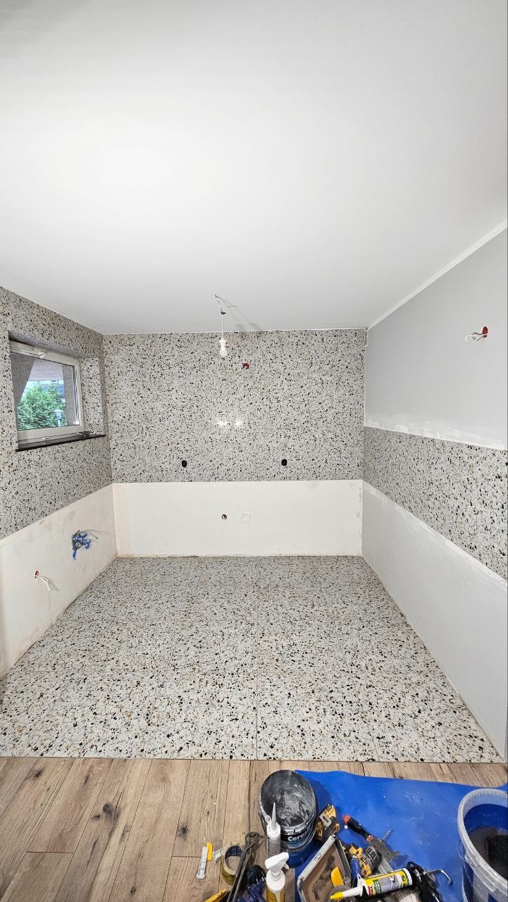 Pokój w trakcie prac wykończeniowych: ściany częściowo pokryte dekoracyjnym tynkiem z efektem kamyczków, dolna partia ścian przygotowana pod malowanie, podłoga wyłożona płytkami imitującymi...