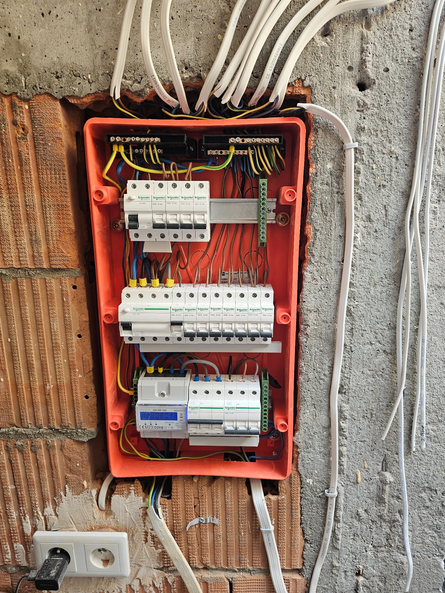 Pomarańczowa skrzynka z bezpiecznikami i licznikiem energii elektrycznej zamontowana na ceglanej ścianie, widoczne kable i gniazdko elektryczne z podłączoną ładowarką.