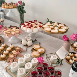 Bogato zastawiony stół z różnorodnymi deserami: mini tartaletki z kremem i owocami, bezy z owocami, puddingi w słoiczkach z owocami, ciastka z napisem 'Mrs & Mrs', dekoracje z różowych kwiatów...