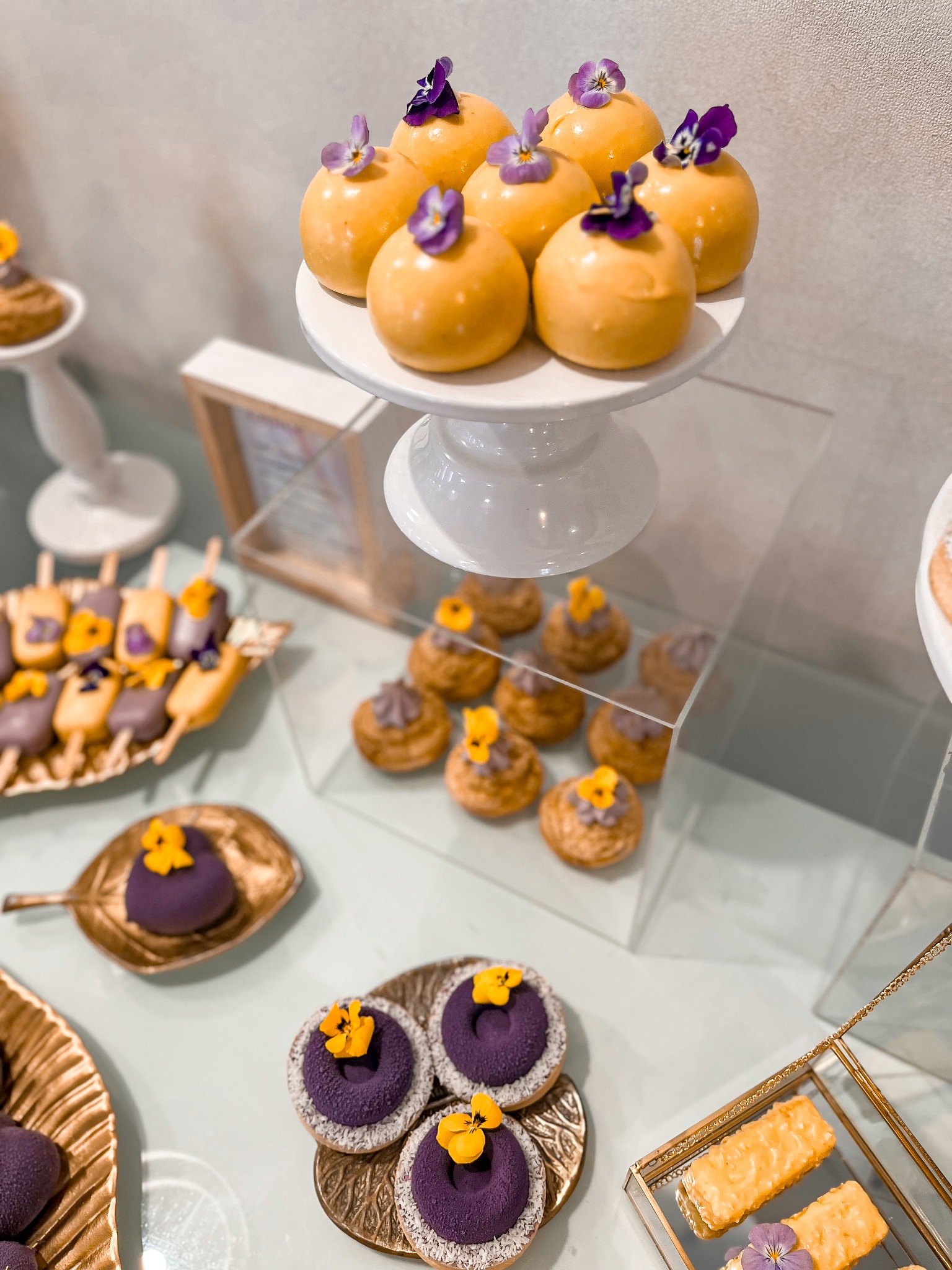 Elegancki stół z różnorodnymi deserami: kulki w polewie z kwiatami, mini bezy z kremem, ciastka na patyku, wszystko w pastelowych kolorach i artystycznej kompozycji.