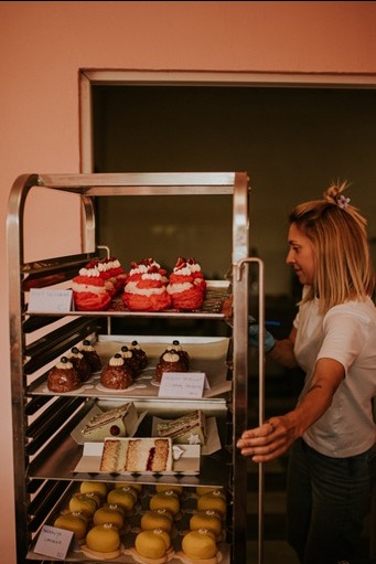 Metalowy regał z czterema półkami wypełnionymi różnorodnymi ciastami i deserami: czerwone babeczki z kremem, czekoladowe mini-tort z borówkami, kawałki tortu z kremem i owocami oraz żółte okrągłe...