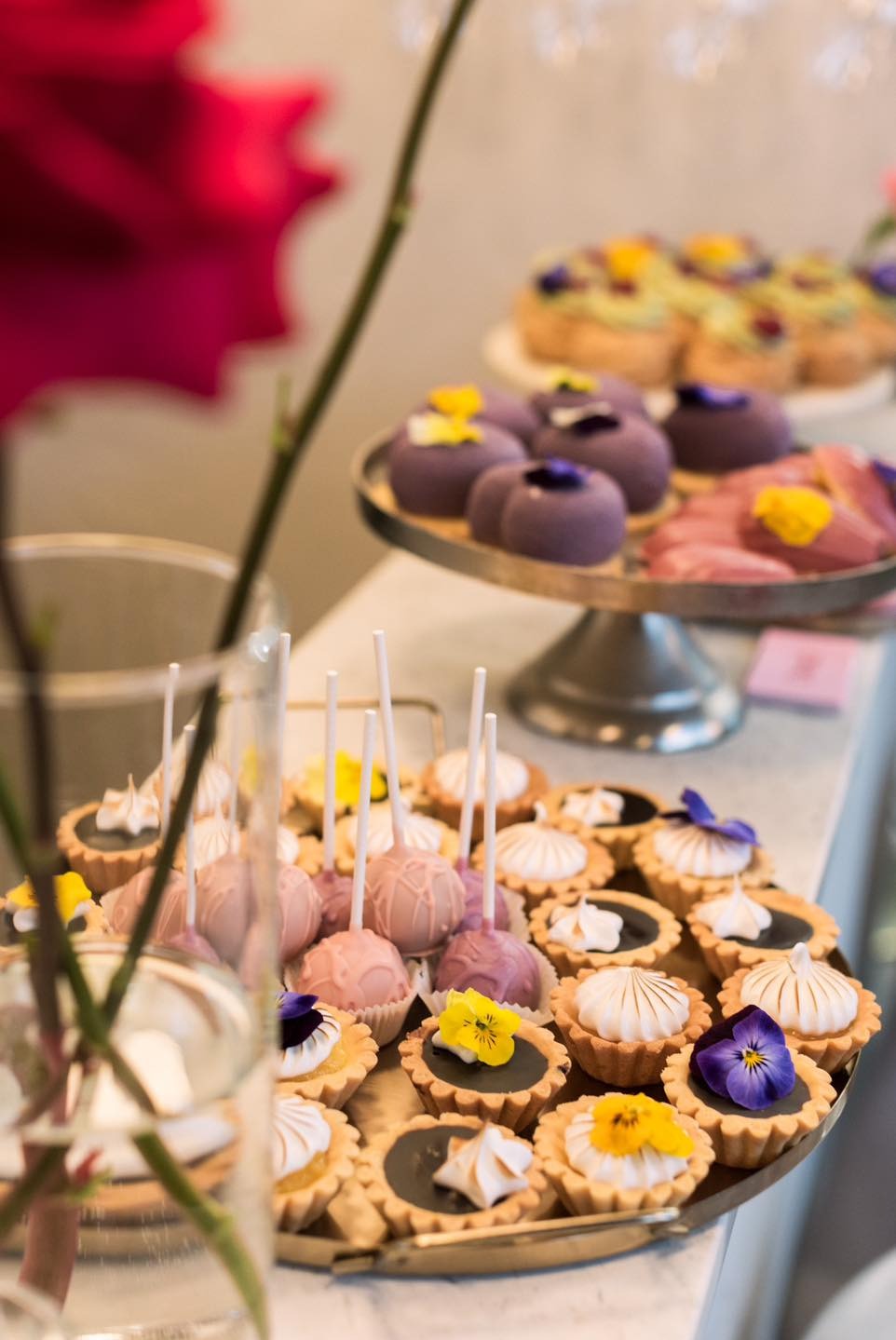 Elegancki zestaw słodkości: mini tartaletki z kremem i kwiatami, cake pops w pastelowych odcieniach oraz deserki w kształcie kopułek na srebrnej paterze, uzupełniony wazonem z czerwoną różą.