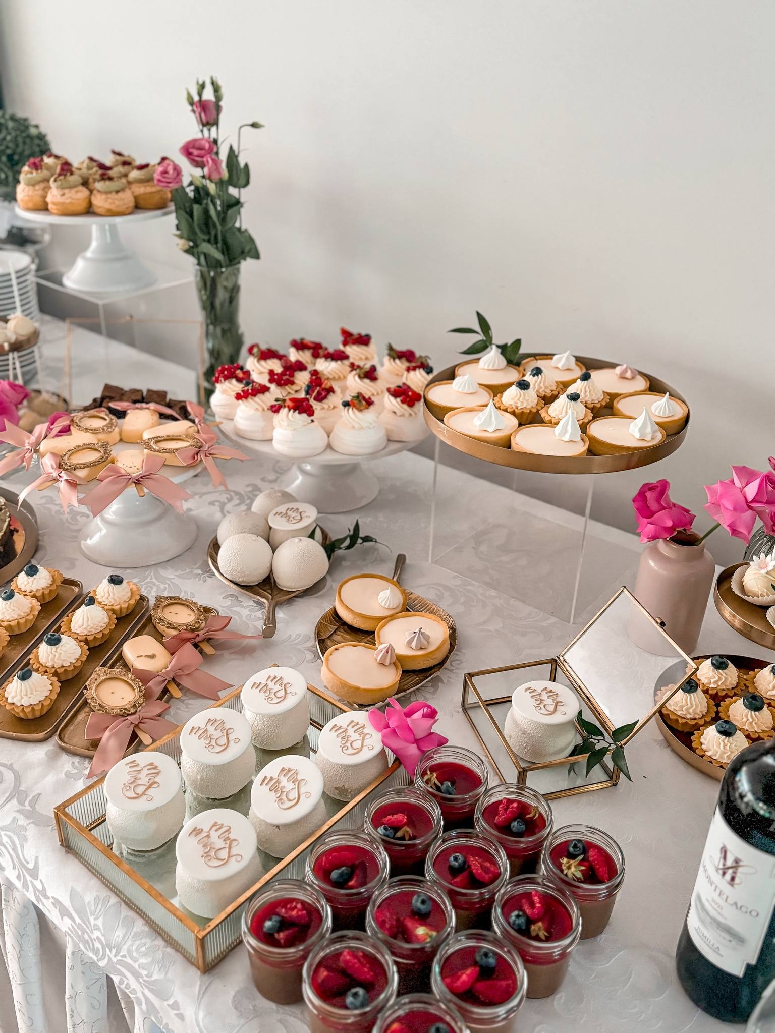 Bogato zastawiony stół z różnorodnymi deserami: mini tartaletki z kremem i owocami, bezy z owocami, puddingi w słoiczkach z owocami, ciastka z napisem 'Mrs & Mrs', dekoracje z różowych kwiatów...