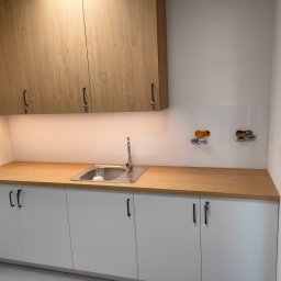 WoodenArt by Dominik Bichta - Nowoczesna kuchnia: białe szafki z czarnymi uchwytami, drewniany blat, stalowy zlew i górne szafki z okleiną drewnopodobną, widoczne gniazdka elektryczne bez osprzętu.