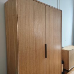 WoodenArt by Dominik Bichta - Dwudrzwiowa szafa z jasnego drewna z zaokrąglonymi krawędziami i czarnymi uchwytami, stojąca na parkiecie w geometryczny wzór, w tle jasna ściana z dekoracyjnymi listwami.