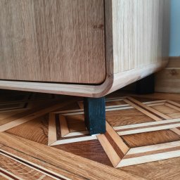 WoodenArt by Dominik Bichta - Fragment szafki z jasnego drewna z zaokrąglonymi krawędziami, wspartej na czarnych, kwadratowych nóżkach, ustawionej na parkiecie w geometryczny wzór.