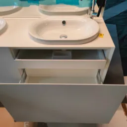 Biała szafka łazienkowa z dwiema umywalkami i wysuniętą szufladą, widoczne elementy montażowe szuflady, w tle zabezpieczone folią lustro i ciemna tkanina.