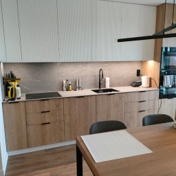 WoodenArt by Dominik Bichta - Nowoczesna kuchnia z drewnianymi frontami szafek dolnych i białymi, ryflowanymi szafkami górnymi, czarny zlew i bateria, marmurowy backsplash, drewniany stół z szarymi krzesłami w tle, widoczne...