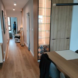 WoodenArt by Dominik Bichta - Nowoczesna zabudowa w przedpokoju z szafą z drewnopodobnymi frontami, przeszkloną witryną z podświetleniem LED i półkami na wino, widok na korytarz z osobą rozmawiającą przez telefon i sprzętem...