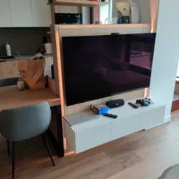 Telewizor wbudowany w drewnianą zabudowę z półkami, z oświetleniem LED, pod nim biała szafka RTV z ryflowanymi frontami, na szafce gry, głośnik i pady do konsoli, obok stół z szarym krzesłem.