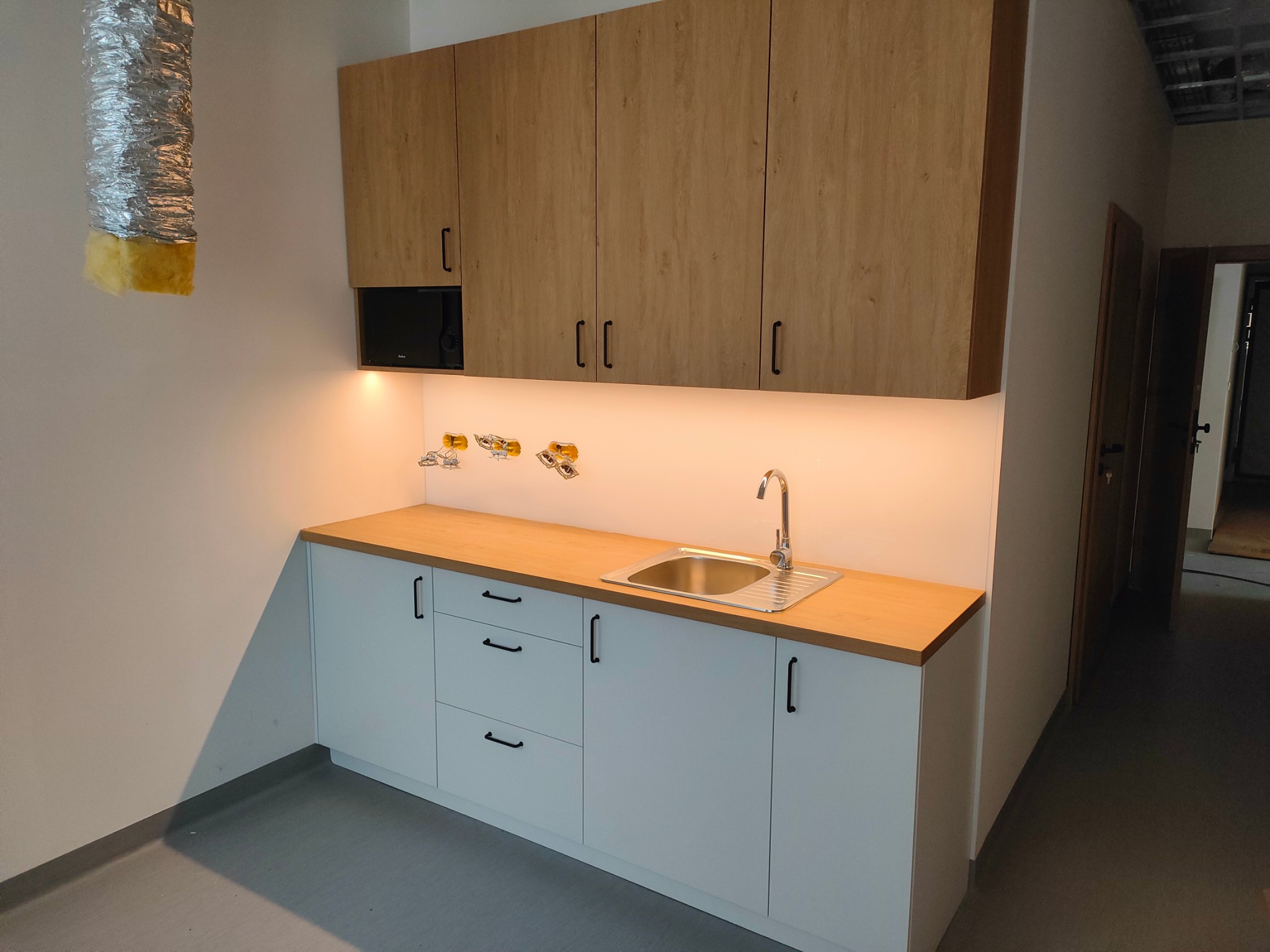 Nowoczesna zabudowa kuchenna z jasnymi, drewnopodobnymi szafkami górnymi i białymi szafkami dolnymi z czarnymi uchwytami, oświetleniem LED pod szafkami, widoczna instalacja wodna nad blatem, w tle...