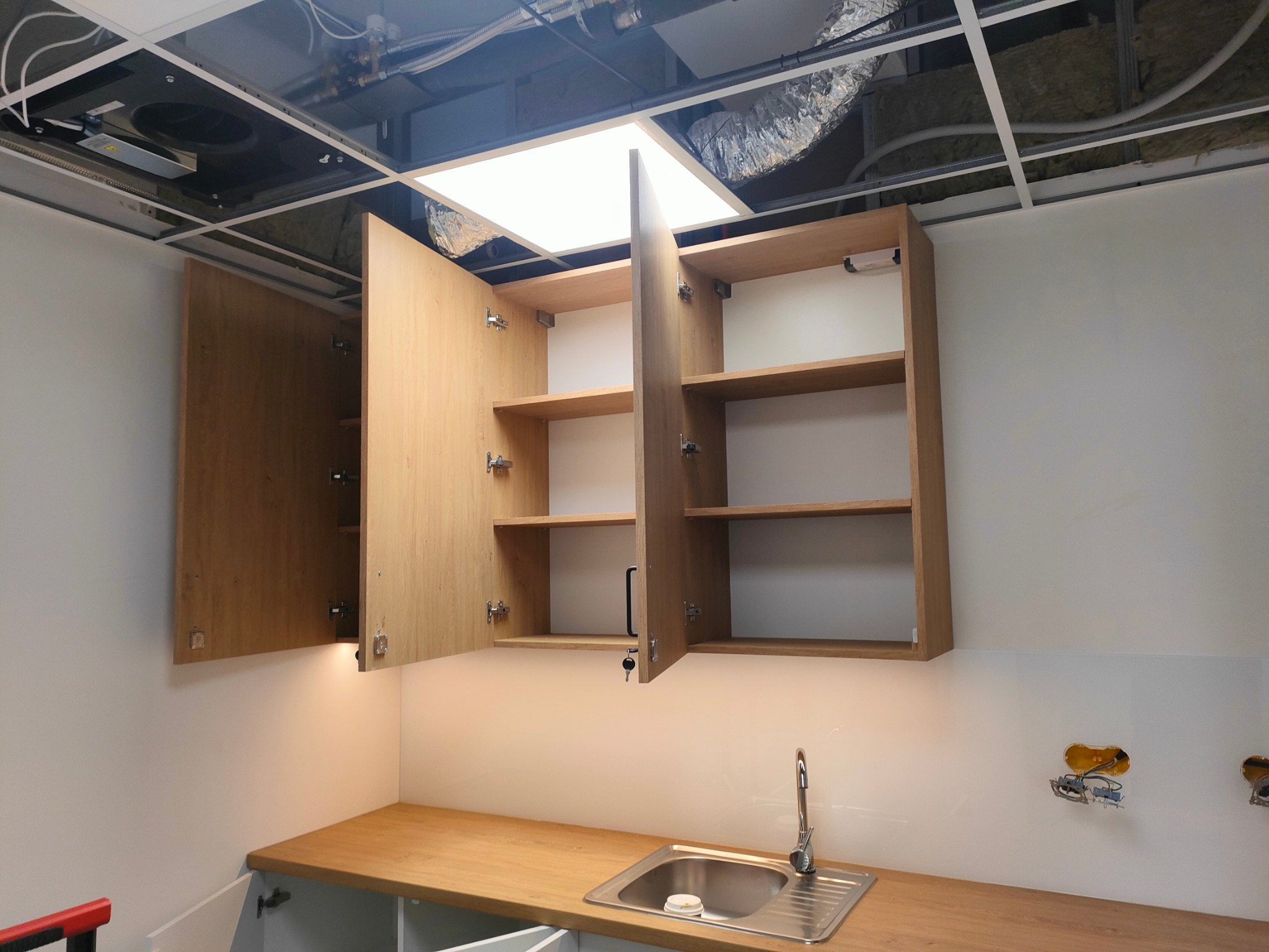 Kuchnia w trakcie montażu: otwarte drewniane szafki wiszące nad blatem z wbudowanym zlewozmywakiem, widoczne niezabezpieczone gniazdka elektryczne na ścianie i sufit podwieszany z oświetleniem.