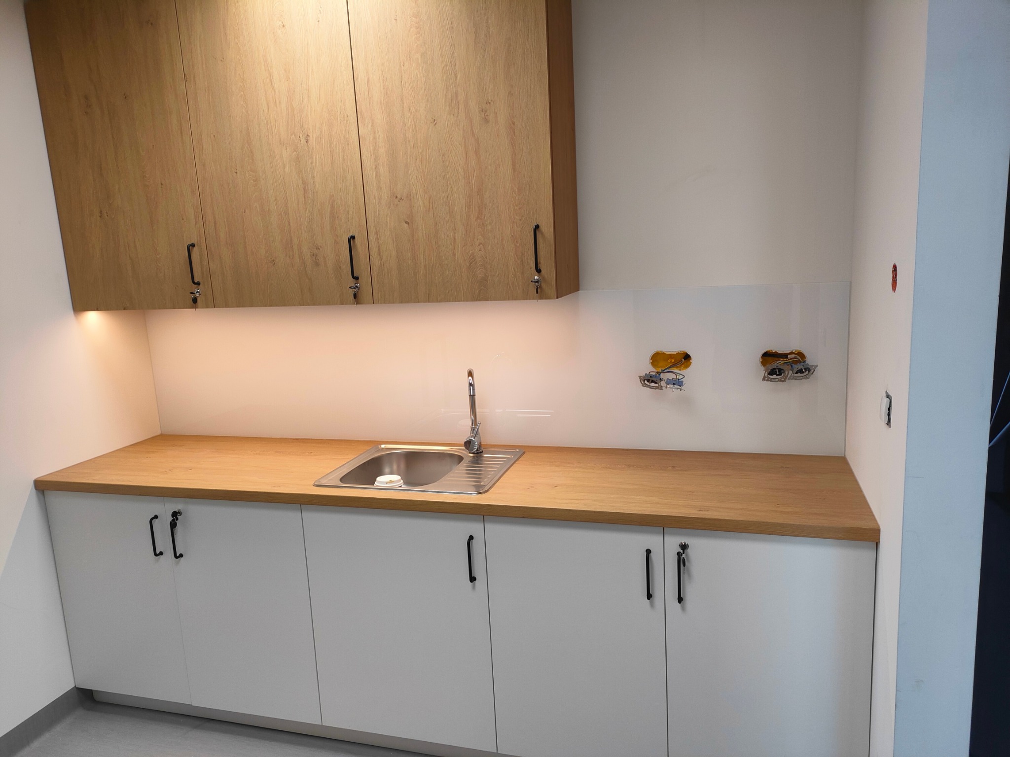 Nowoczesna kuchnia: białe szafki z czarnymi uchwytami, drewniany blat, stalowy zlew i górne szafki z okleiną drewnopodobną, widoczne gniazdka elektryczne bez osprzętu.