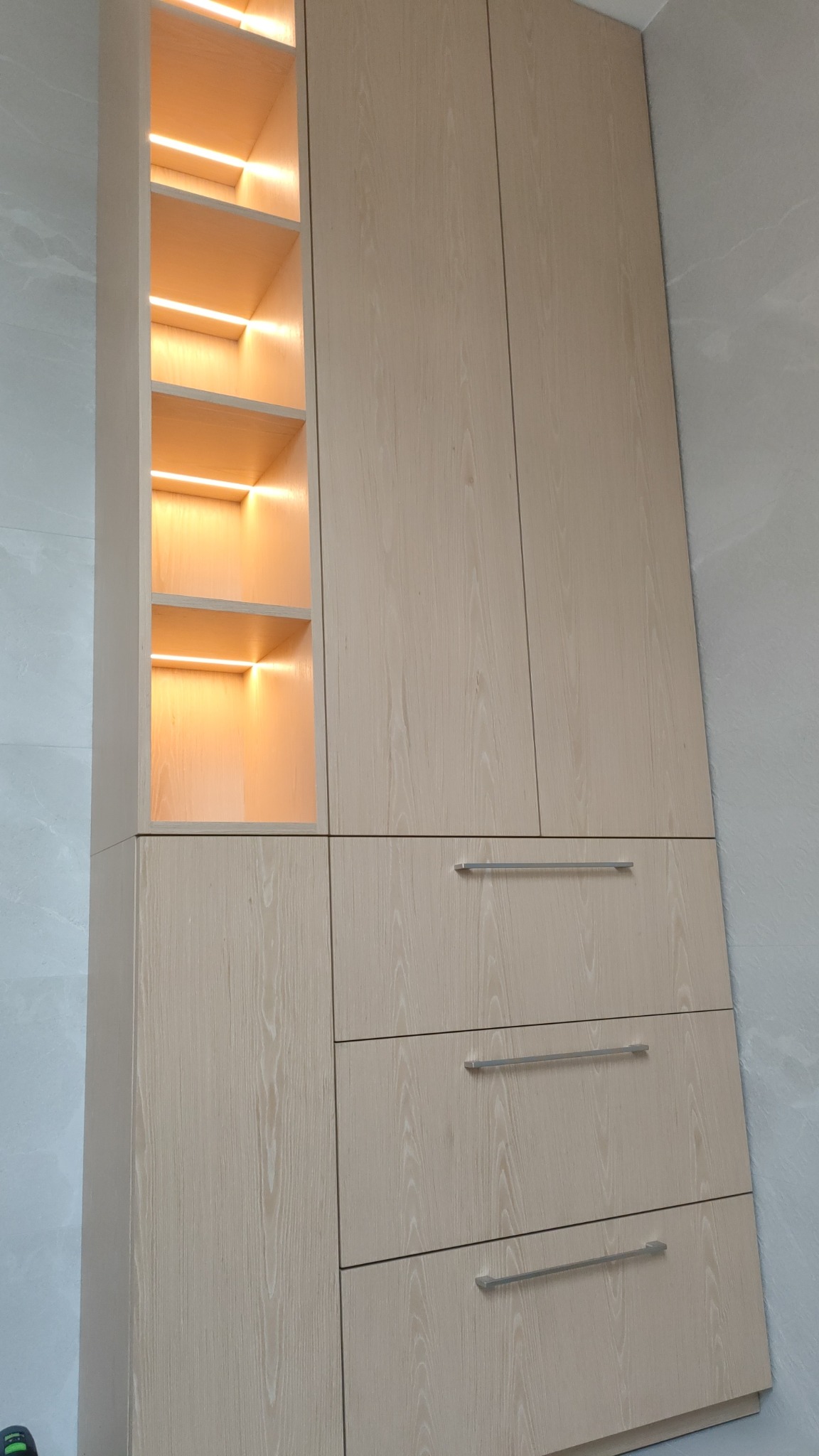 Jasna, drewnopodobna szafa wnękowa z oświetleniem LED w otwartych półkach, z trzema szufladami z metalowymi uchwytami.