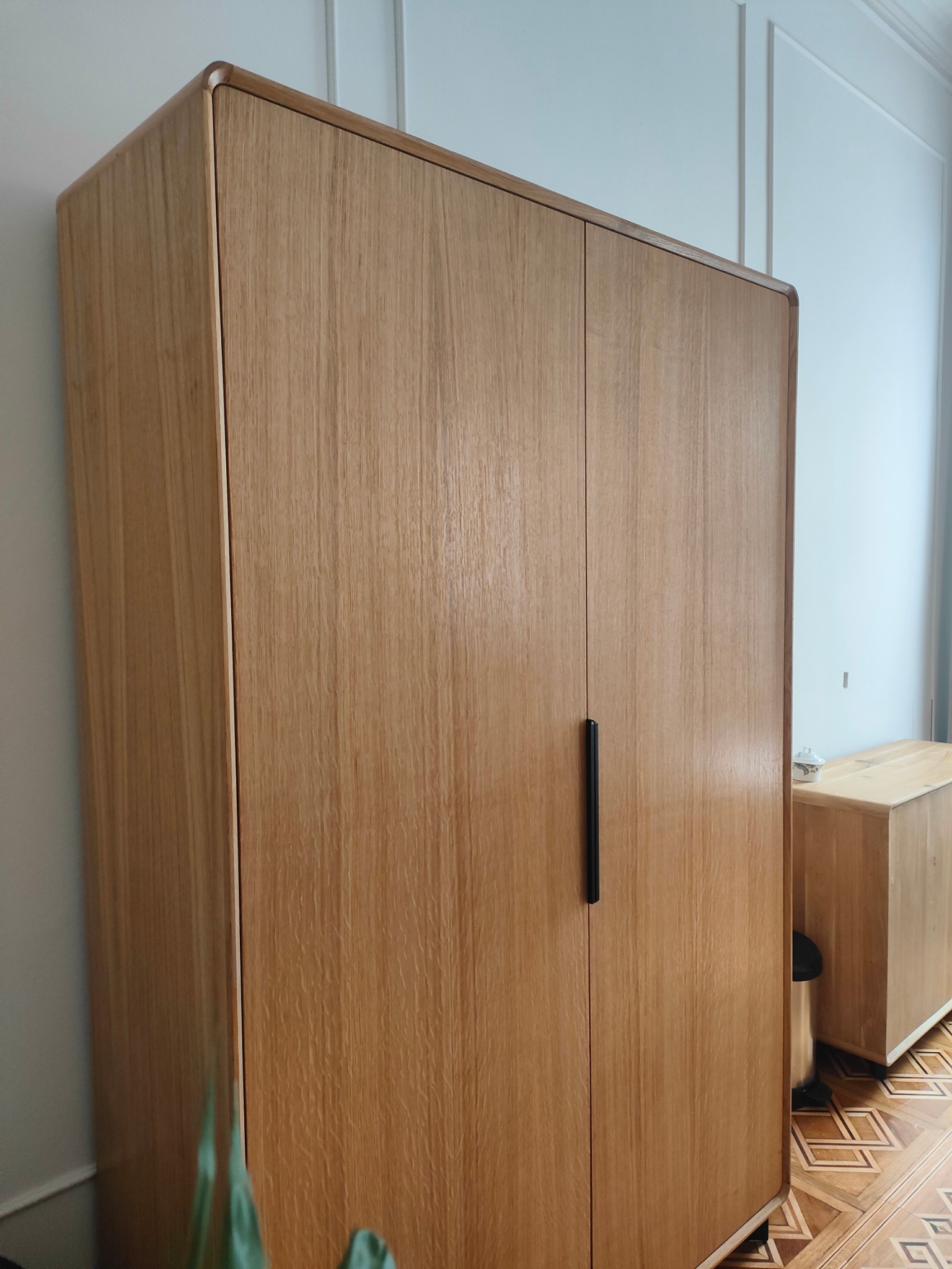 Dwudrzwiowa szafa z jasnego drewna z zaokrąglonymi krawędziami i czarnymi uchwytami, stojąca na parkiecie w geometryczny wzór, w tle jasna ściana z dekoracyjnymi listwami.