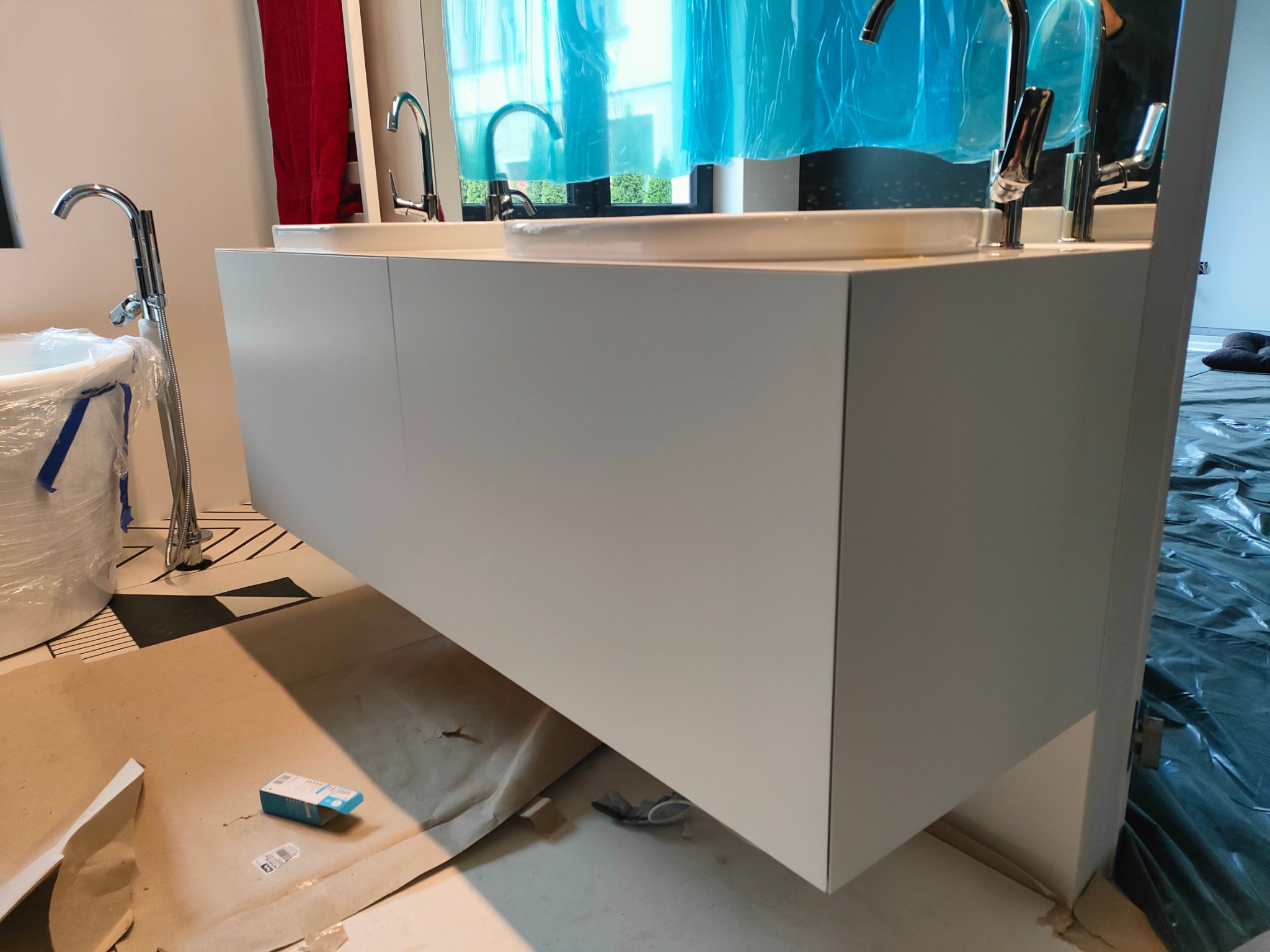 Biała szafka pod umywalkę z dwiema umywalkami i chromowanymi kranami, podłoga częściowo zabezpieczona folią malarską, widoczne akcesoria montażowe.