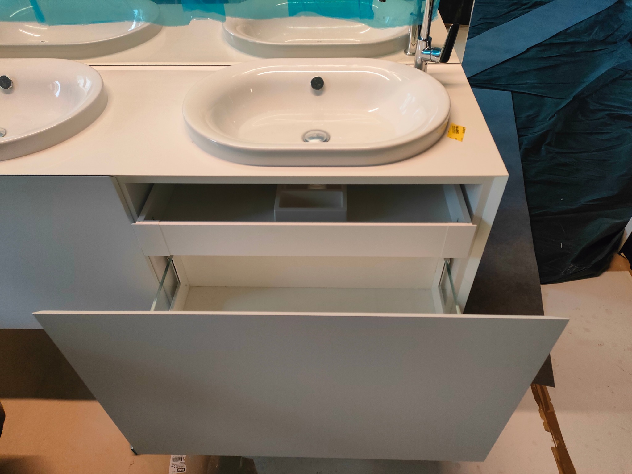 Biała szafka łazienkowa z dwiema umywalkami i wysuniętą szufladą, widoczne elementy montażowe szuflady, w tle zabezpieczone folią lustro i ciemna tkanina.