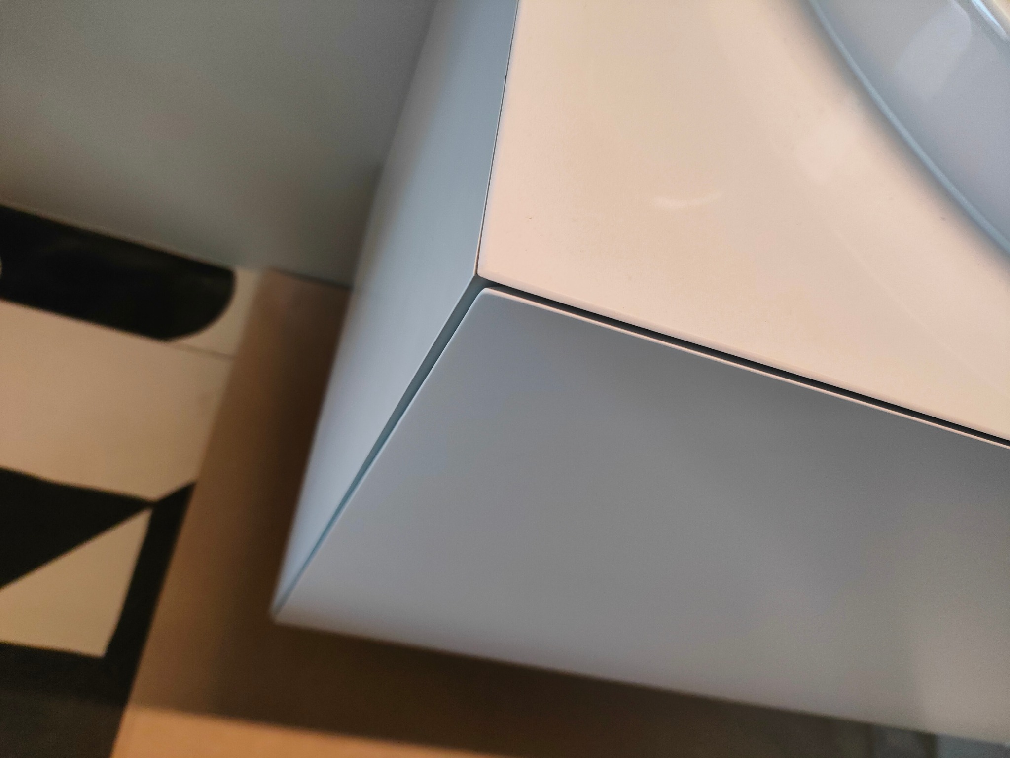 Fragment białej szafki łazienkowej z umywalką, widoczny z dołu pod kątem, na tle wzorzystej podłogi.