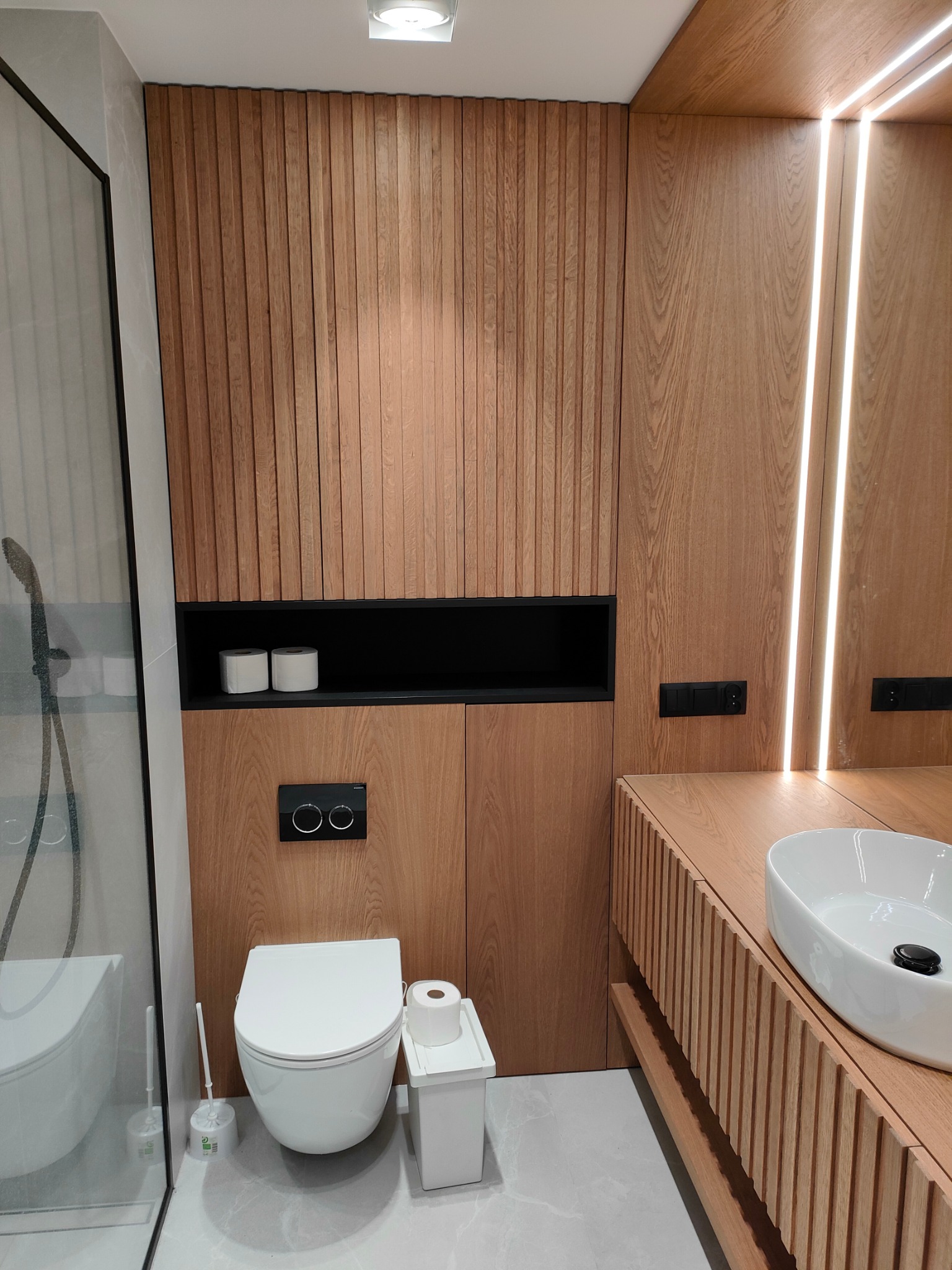 Nowoczesna łazienka z drewnianą zabudową: pionowe lamele z jasnego drewna na ścianie i szafce pod umywalką, podświetlenie LED, wisząca toaleta.