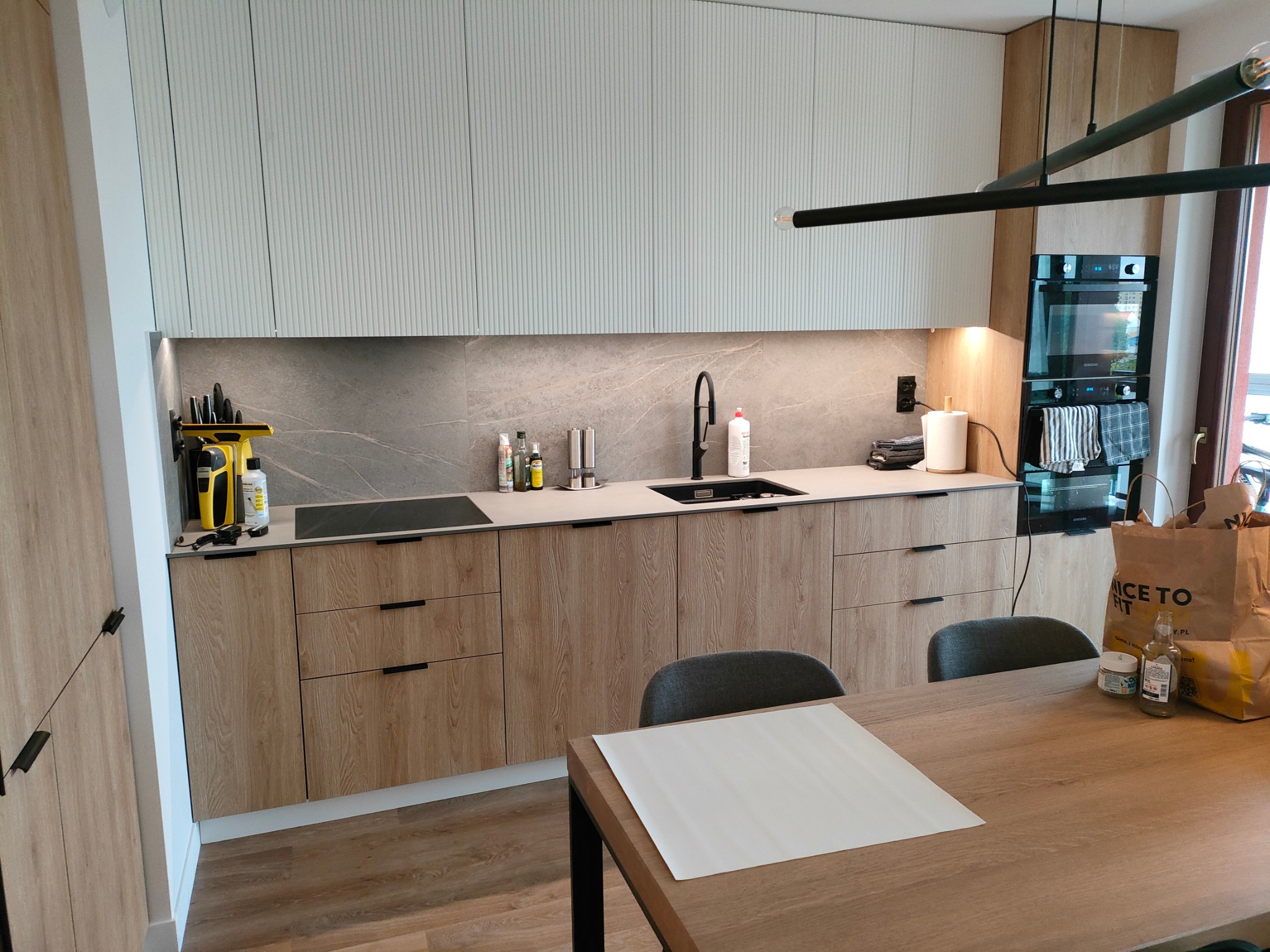 Nowoczesna kuchnia z drewnianymi frontami szafek dolnych i białymi, ryflowanymi szafkami górnymi, czarny zlew i bateria, marmurowy backsplash, drewniany stół z szarymi krzesłami w tle, widoczne...