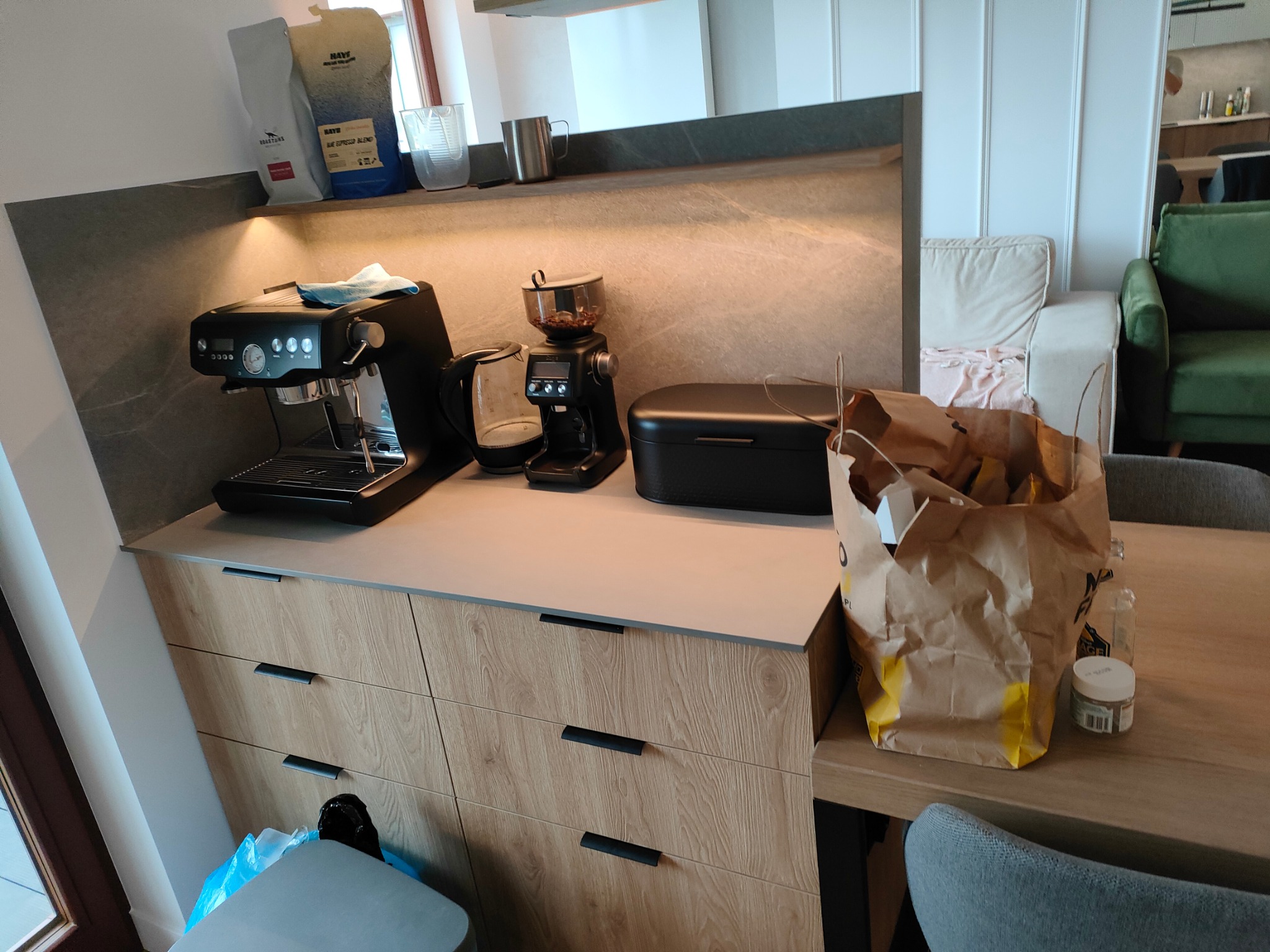 Nowoczesna kuchnia z drewnianymi szafkami, blatem w kolorze szarości i sprzętem do kawy: ekspres, młynek i czajnik elektryczny. Na blacie stoi torba papierowa z zakupami.