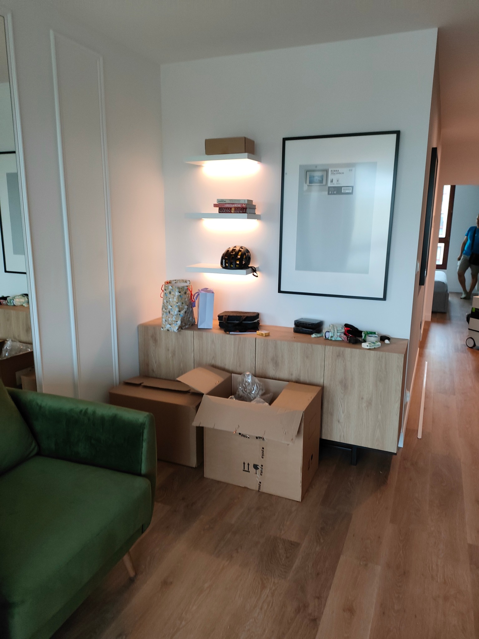 Jasna komoda z drewnianymi frontami, na niej ozdoby i przedmioty, nad nią półki z podświetleniem, obok zielony fotel i kartony, w tle osoba w przejściu.