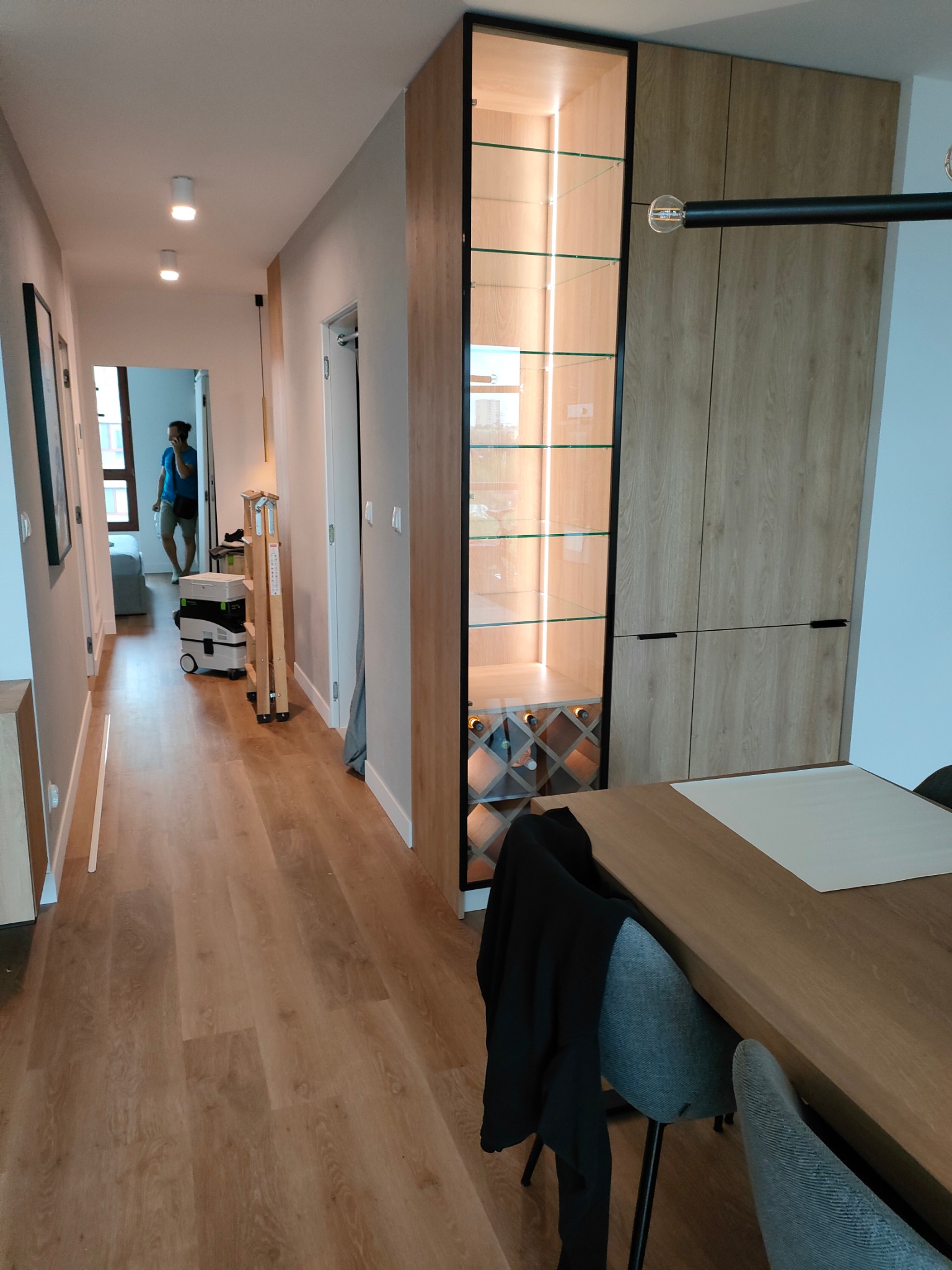 Nowoczesna zabudowa w przedpokoju z szafą z drewnopodobnymi frontami, przeszkloną witryną z podświetleniem LED i półkami na wino, widok na korytarz z osobą rozmawiającą przez telefon i sprzętem...