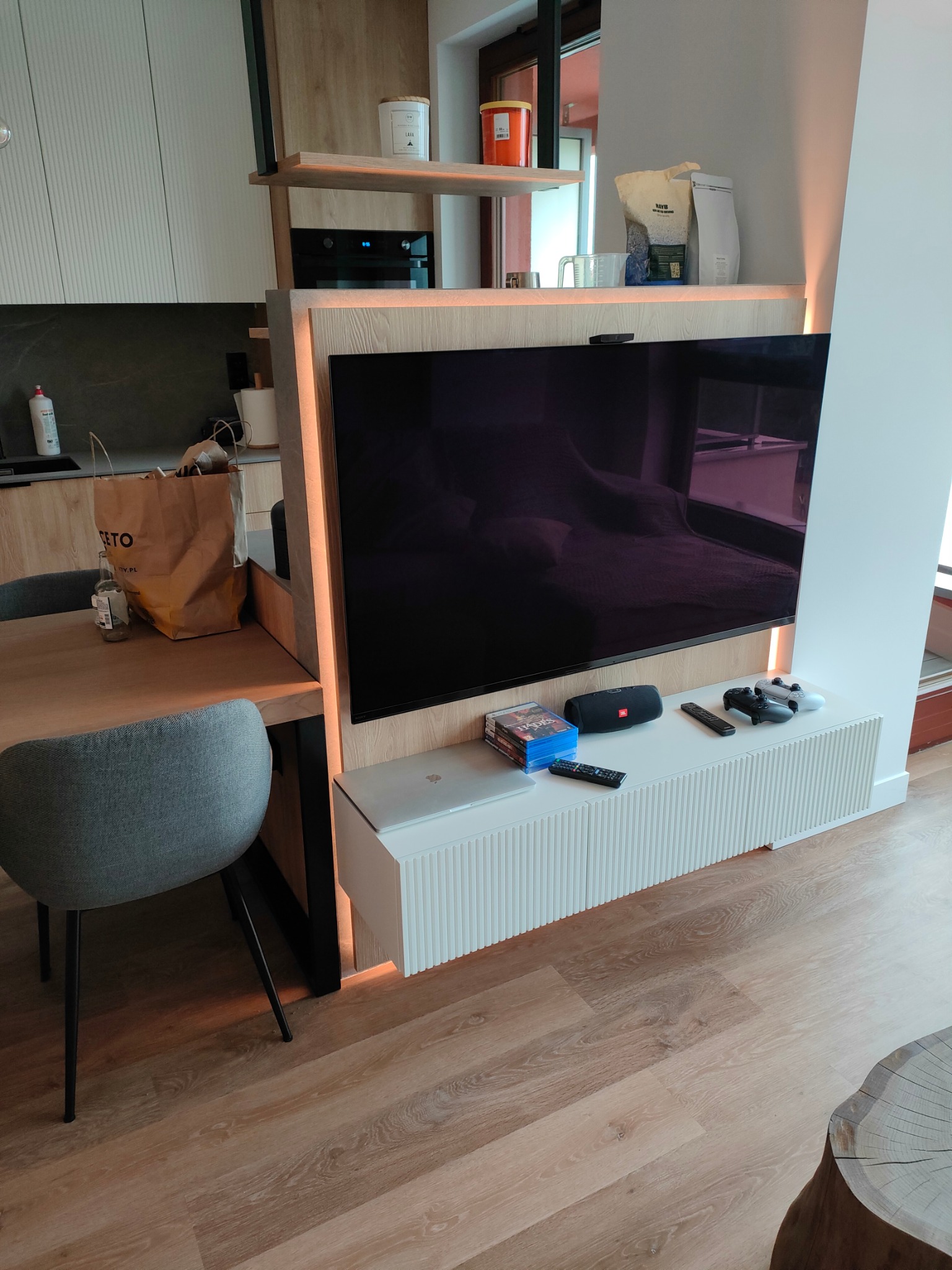 Telewizor wbudowany w drewnianą zabudowę z półkami, z oświetleniem LED, pod nim biała szafka RTV z ryflowanymi frontami, na szafce gry, głośnik i pady do konsoli, obok stół z szarym krzesłem.
