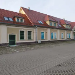 Długi, parterowy budynek z czerwonym dachem i beżową elewacją, okna w zielonych ramach, widok z zewnątrz na pusty parking z betonowej kostki.