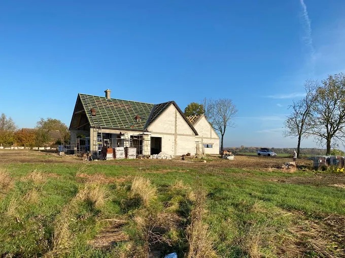 Dom w trakcie budowy, widoczne ściany w stanie surowym, więźba dachowa z zieloną folią, rusztowania i materiały budowlane na posesji w słoneczny dzień.